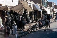 The bustling sidewalks of Kandahar with its open-fronted shops. Although very much a man's world, a group of women is walking past. Donkey transport is very  common.